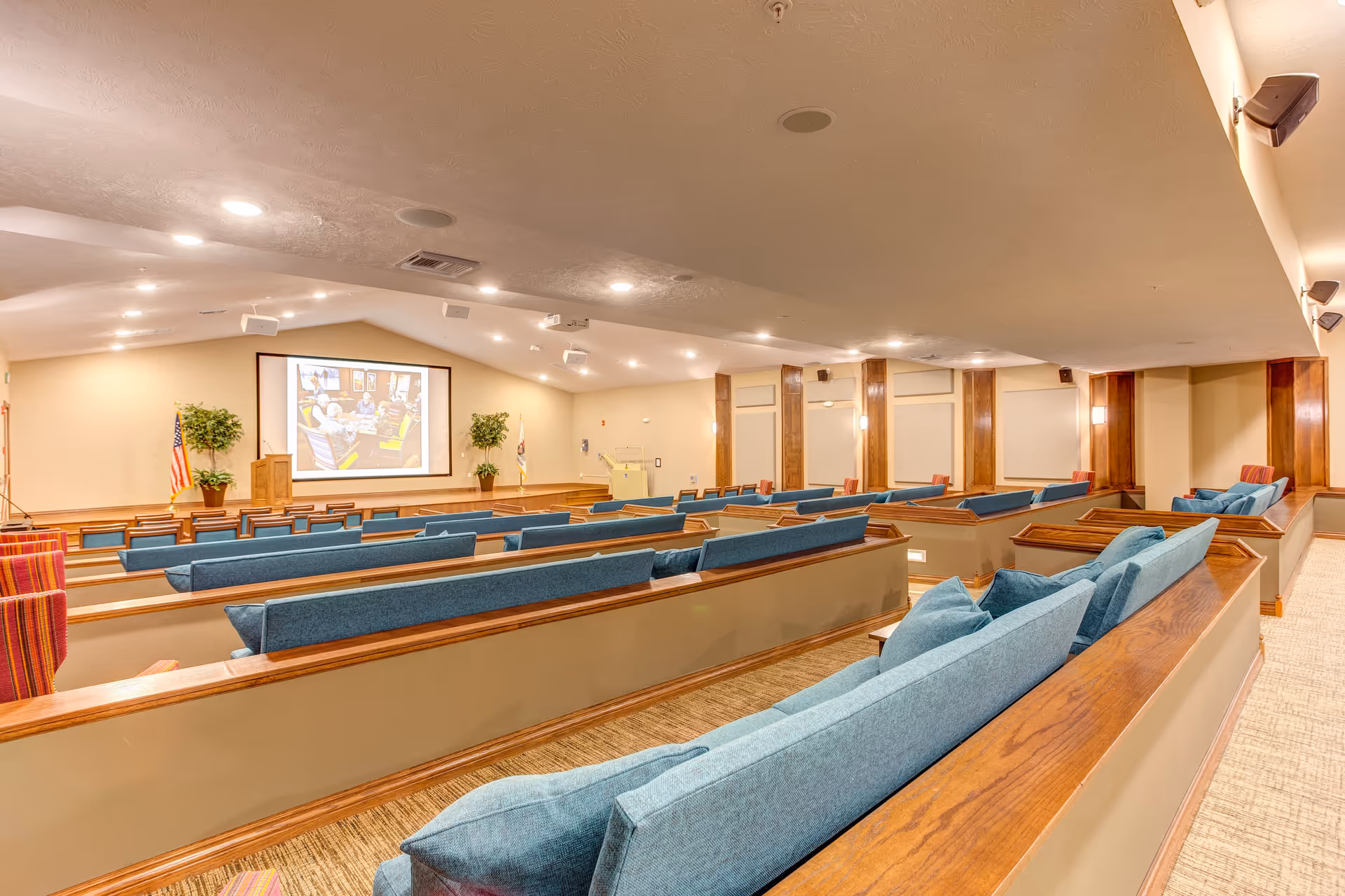 A spacious auditorium or presentation room with tiered seating featuring blue cushioned benches and wooden armrests. At the front, there is a large projection screen displaying an image, a wooden podium, and two potted plants on either side. The room has beige walls, recessed lighting in the ceiling, and wall-mounted speakers.