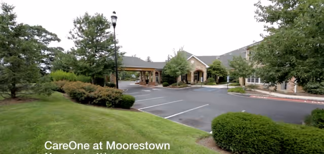 Exterior view of CareOne at Moorestown facility showing a parking lot, landscaped greenery, trees, and the entrance to the building with a covered drop-off area.