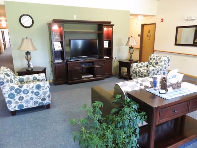 Seating area with a large wooden TV entertainment center, patterned armchairs, side tables with lamps, and a console table with decor.