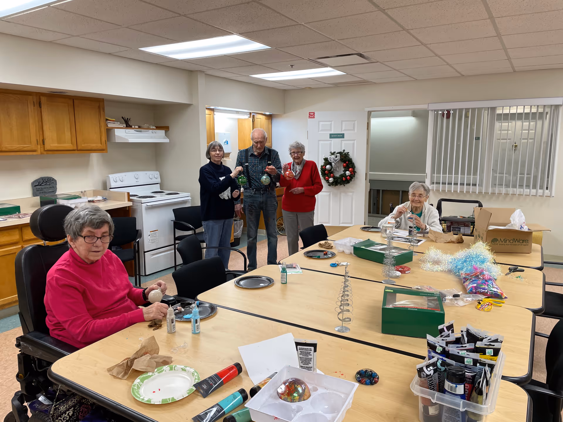 A group of elderly individuals engaged in a craft activity in a room with a kitchen area. Four people are standing near the back holding decorated ornaments, while two others are seated at a table working on crafts. The table is covered with art supplies, including paint tubes, brushes, and decorative materials. The room has wooden cabinets, a white stove, and a door decorated with a holiday wreath.