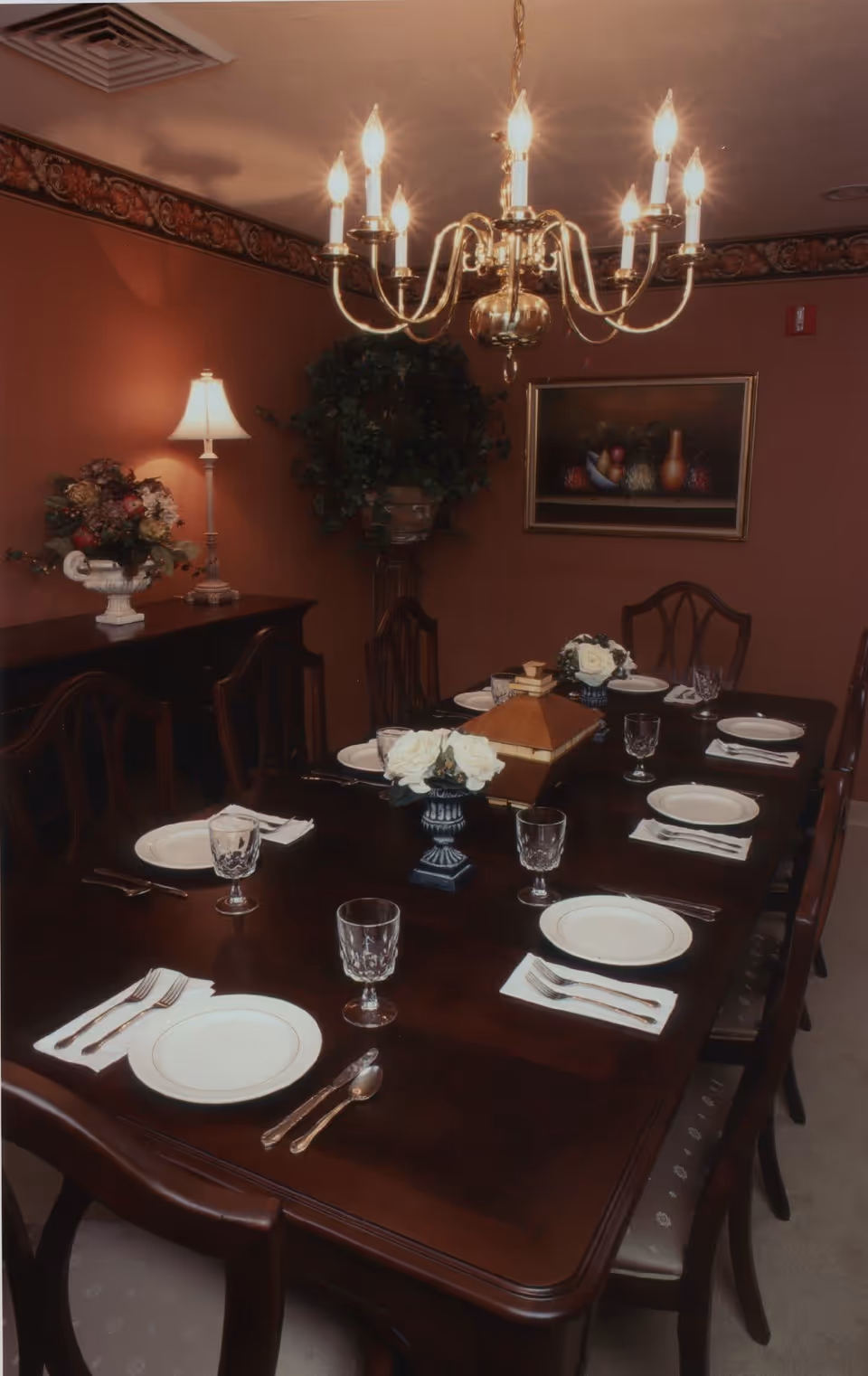 A formal dining room with a dark wooden table set for eight people. The table is arranged with white plates, silverware, crystal glasses, and white napkins. Two small floral centerpieces and a decorative wooden box are placed on the table. The room has warm brown walls with a floral border near the ceiling, a chandelier with lit bulbs hanging above the table, a sideboard with a lamp and floral arrangement, and a framed painting on the wall.