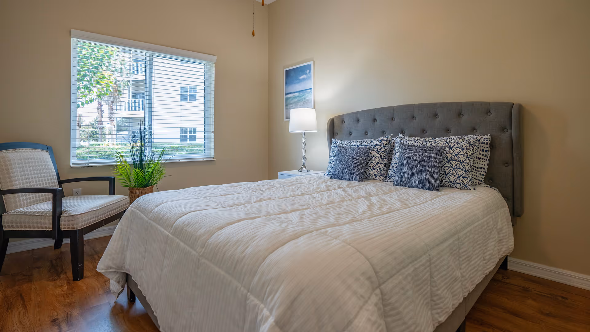A bedroom with a large bed featuring a tufted gray headboard, white bedding, and four decorative pillows in blue and white patterns. To the left of the bed is a window with white blinds, a potted plant, and a cushioned chair with dark wooden arms and legs. A nightstand with a lamp and a framed picture of a beach scene are on the right side of the bed. The floor is wooden and the walls are painted beige.
