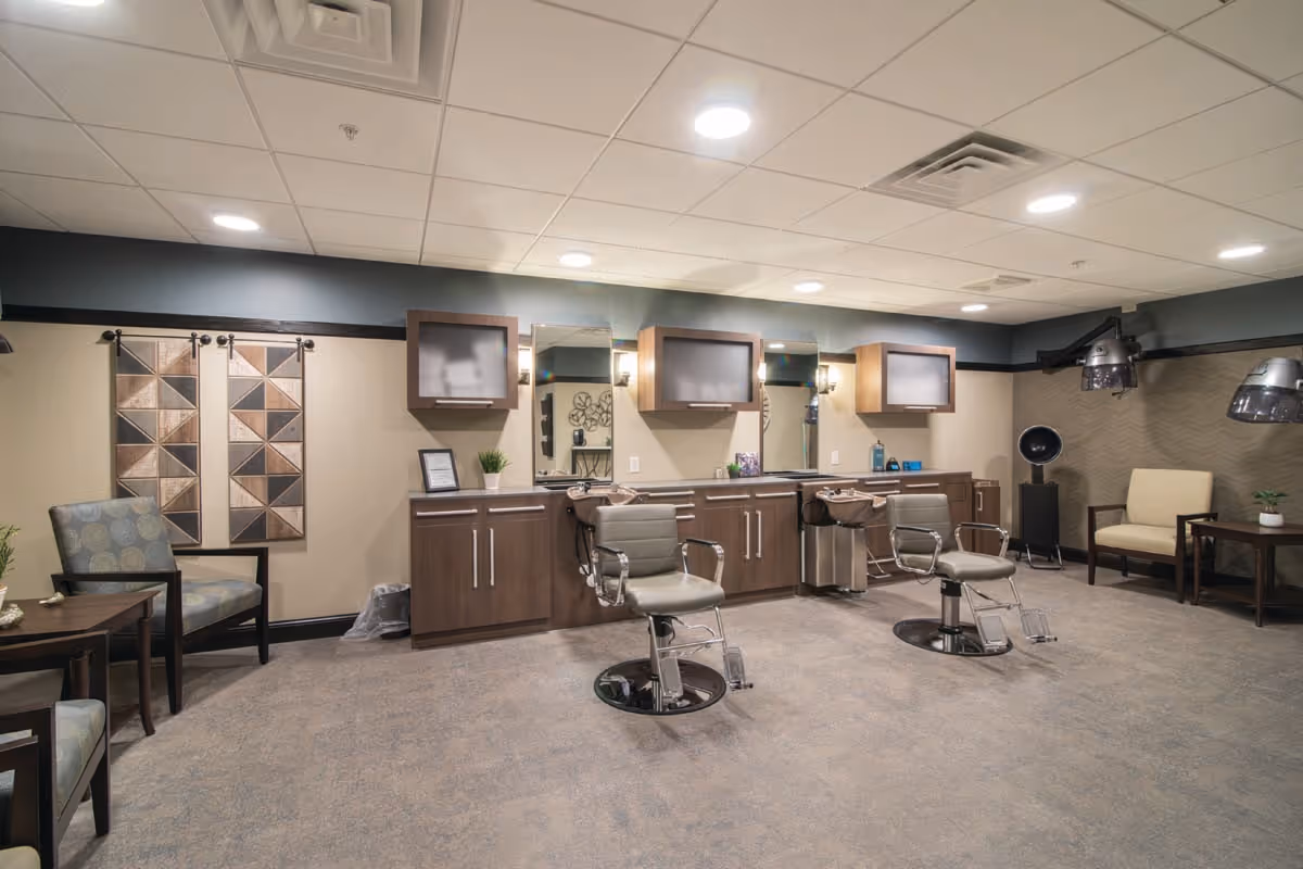 Interior view of a salon area in Orchard Path facility featuring two salon chairs in front of sinks and mirrors, wooden cabinets with frosted glass doors, additional seating with armchairs and side tables, and hair drying equipment mounted on the walls.