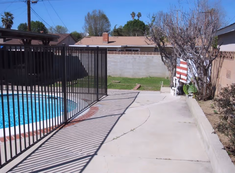 Backyard showing a fenced swimming pool and concrete walkway with fence shadows, a cinderblock privacy wall and houses in the background.