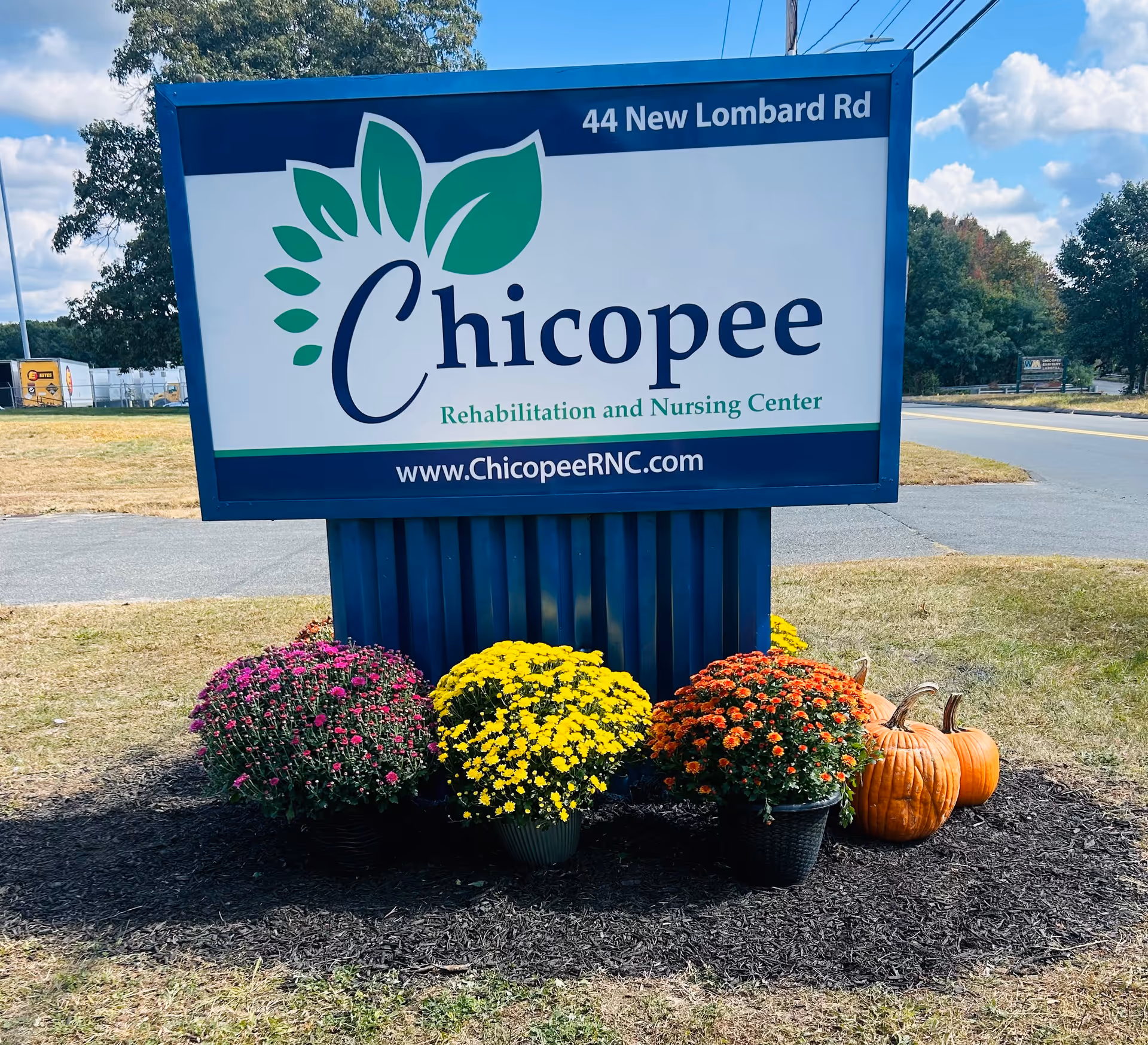Outdoor sign for Chicopee Rehabilitation and Nursing Center at 44 New Lombard Rd, with colorful flowers and pumpkins arranged at the base of the sign, set against a backdrop of trees and a road.