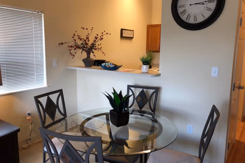 A small dining area with a round glass table and four metal chairs with cushioned seats. On the table is a small potted plant. Behind the table is a counter with decorative items including a vase with branches and a small plant. A large wall clock and a small framed sign that says 'BEGIN' are visible on the wall. A window with closed blinds is on the left side.