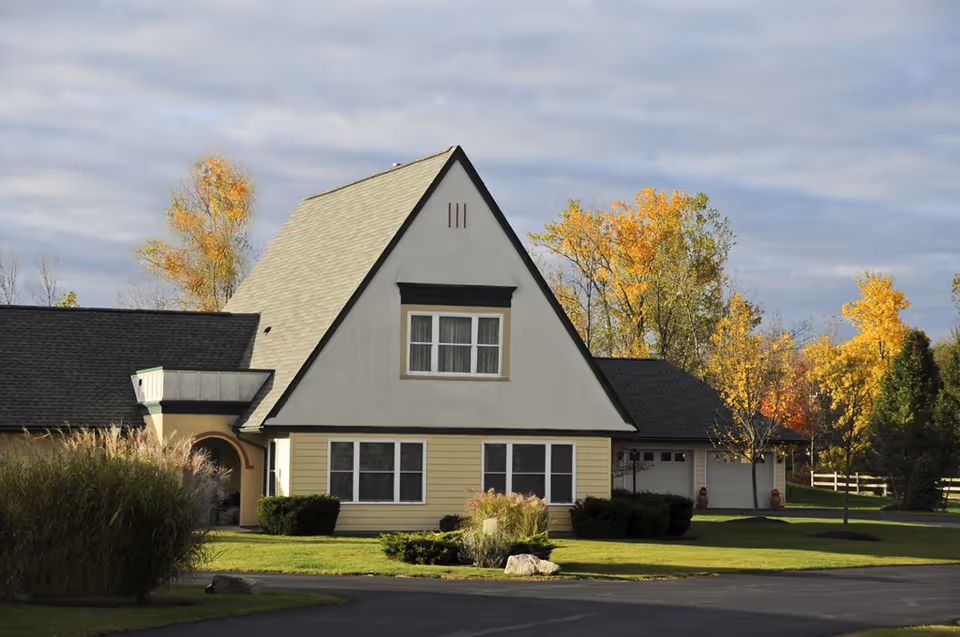 Exterior view of a residential-style building with a steeply pitched roof, surrounded by green lawn and trees with autumn foliage under a cloudy sky.