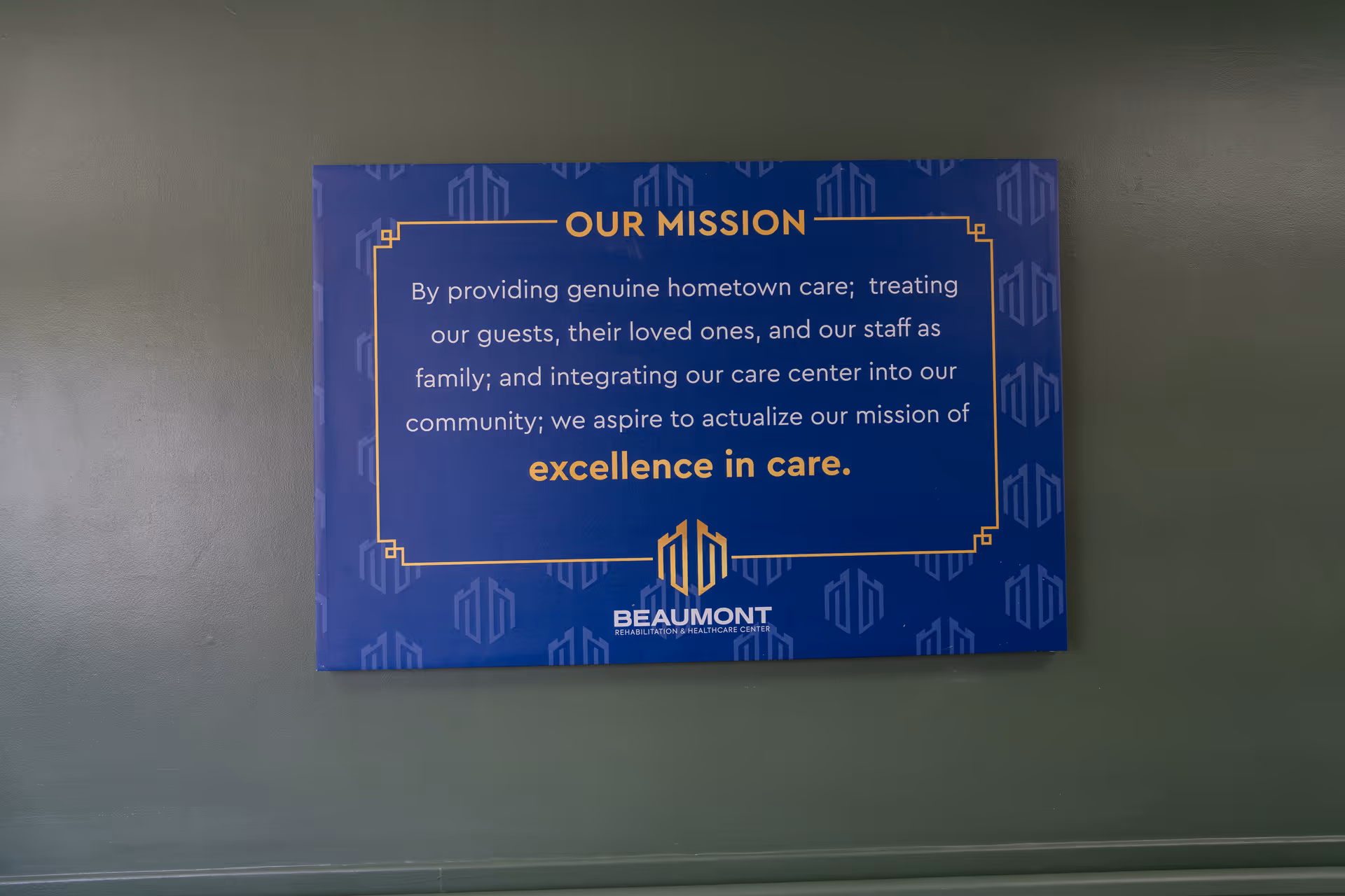 A blue sign mounted on a green wall displaying the mission statement of Beaumont Rehabilitation and Healthcare Center. The sign reads: 'OUR MISSION By providing genuine hometown care; treating our guests, their loved ones, and our staff as family; and integrating our care center into our community; we aspire to actualize our mission of excellence in care.' The Beaumont logo is at the bottom of the sign.