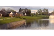 Pond with a fountain in front of a row of single-story cottages and landscaped lawns under a cloudy sky.