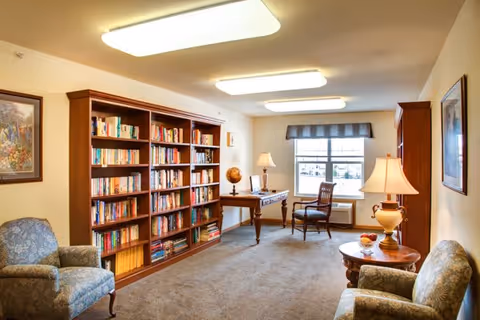 A cozy senior living room with two upholstered armchairs, a wooden bookshelf filled with books, a wooden desk with a globe and lamp, a wooden chair, and a window with a valance letting in natural light. The room is warmly lit with ceiling lights and table lamps, and has carpeted flooring.