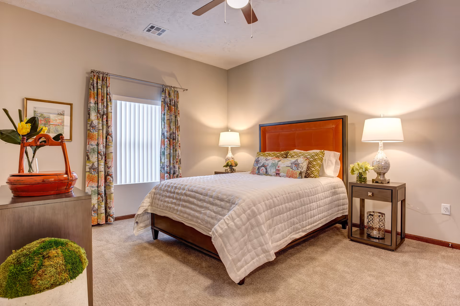A cozy bedroom with a bed featuring a white quilted bedspread and decorative pillows. The bed has a wooden frame with an orange upholstered headboard. There are two bedside tables with lamps and a small flower arrangement on one. A window with vertical blinds and colorful curtains is on the left wall. A dresser with a red decorative basket and a framed picture is also visible. The room has beige walls and carpeted floor.