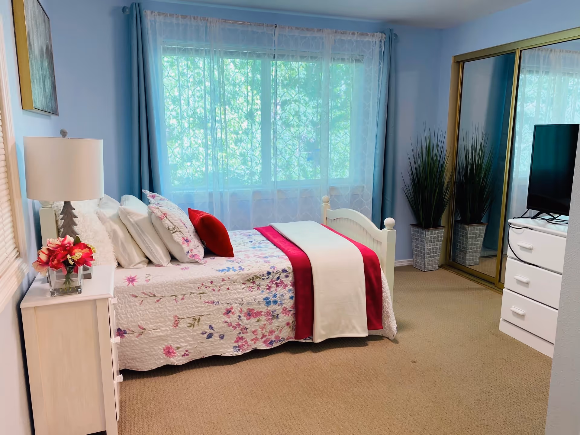 Sunlit bedroom with a floral bedspread, white nightstand and lamp, mirrored closet doors, and a TV on a dresser.