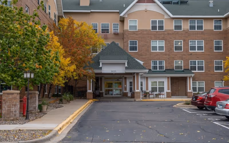 Front exterior view of a multi-story senior living facility building with a covered entrance, several windows, autumn trees with yellow and orange leaves, a sidewalk, and parked cars in the parking lot.