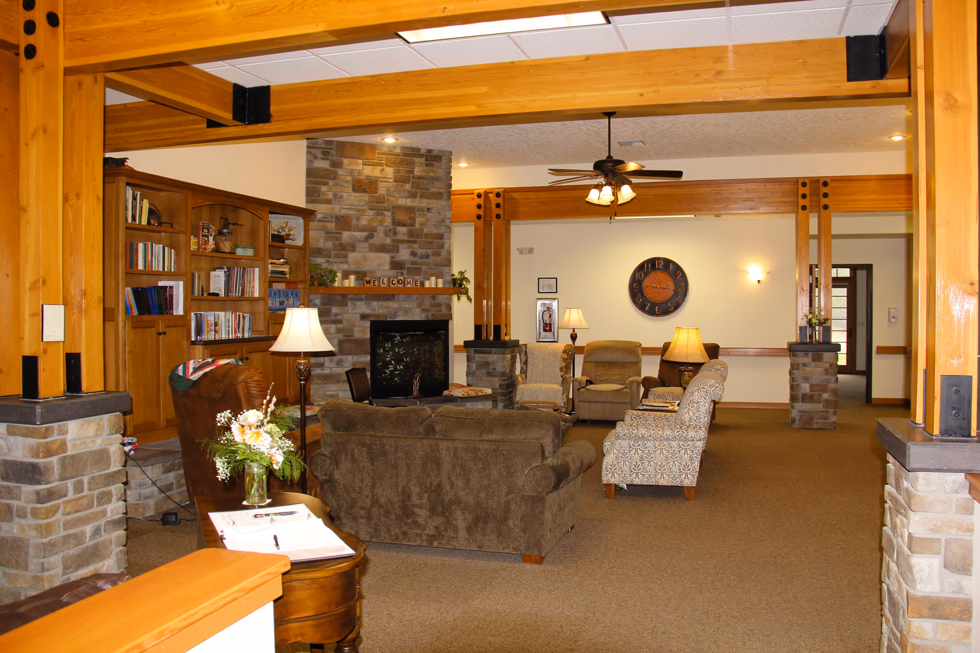 A cozy living room area in an assisted living facility featuring a stone fireplace with a wooden mantle displaying a welcome sign. The room has wooden beams and columns with stone bases, a ceiling fan with lights, a large clock on the wall, several upholstered chairs and sofas arranged for conversation, a wooden bookshelf filled with books, and table lamps providing warm lighting.