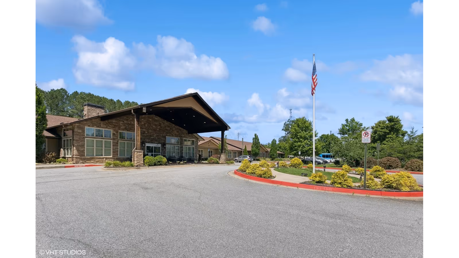 Exterior view of a single-story senior living facility building with a covered entrance, surrounded by landscaped greenery and a flagpole with an American flag. The sky is blue with scattered clouds.
