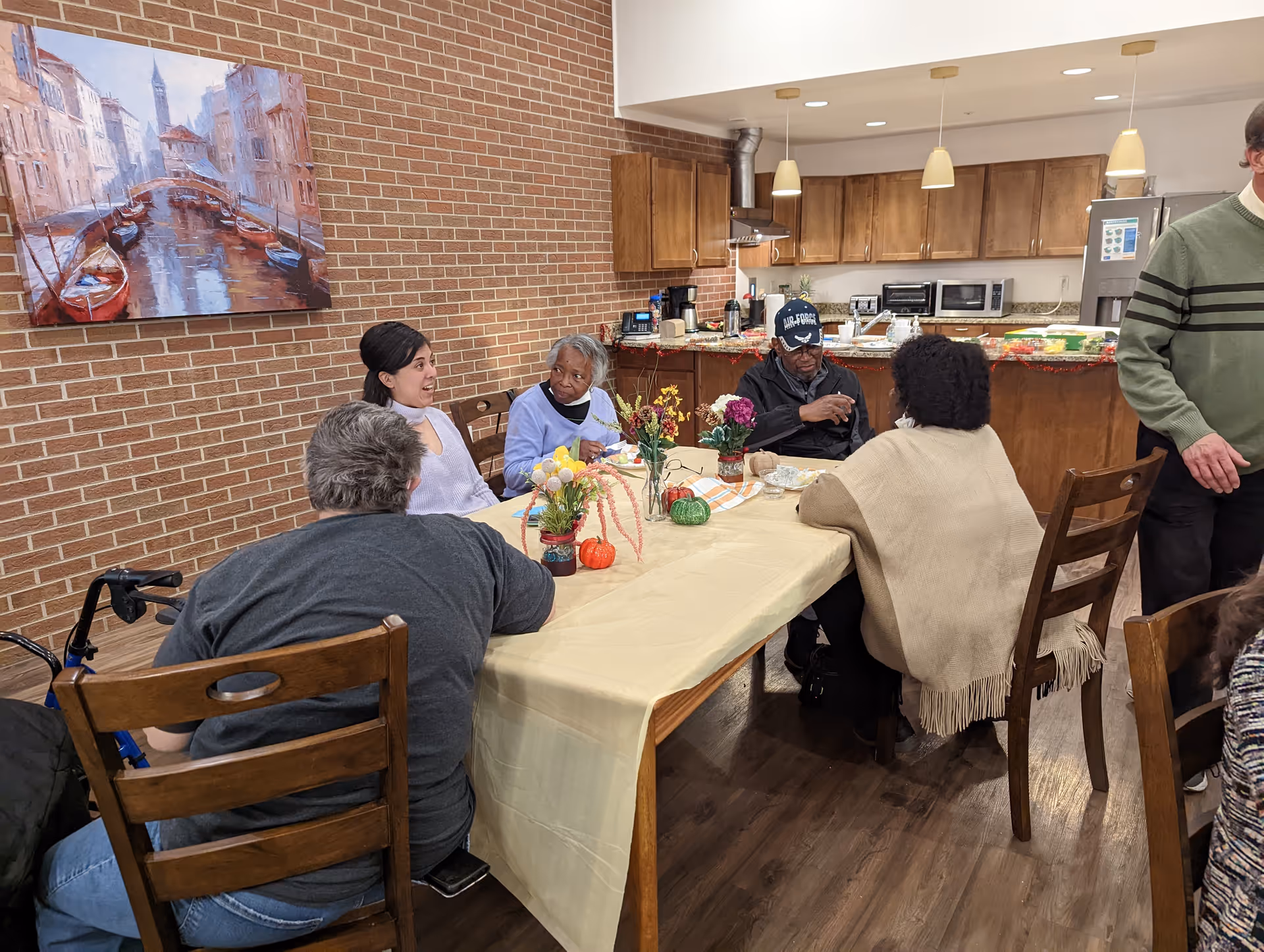 A group of elderly people and a caregiver sitting around a table in a kitchen area, engaging in conversation. The table is covered with a light yellow tablecloth and decorated with small flower arrangements and seasonal decorations. The kitchen in the background has wooden cabinets, a refrigerator, microwave, and other appliances. A painting of a canal scene hangs on the brick wall.