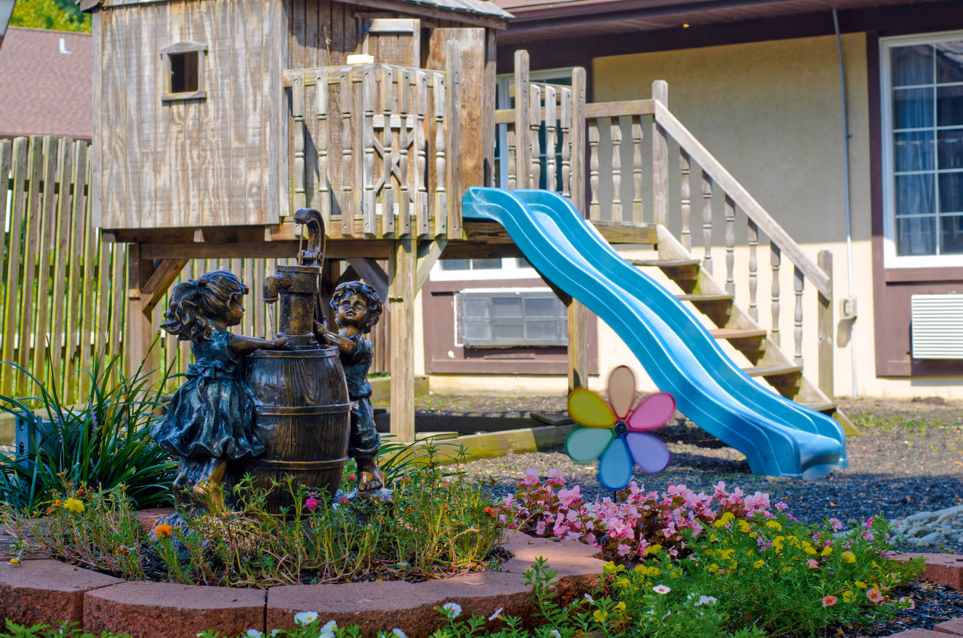 Outdoor playground area with a wooden playhouse on stilts, a blue double slide, and a colorful pinwheel in a garden bed with flowers. In the foreground, there is a bronze statue of two children playing with a water pump surrounded by plants and flowers.