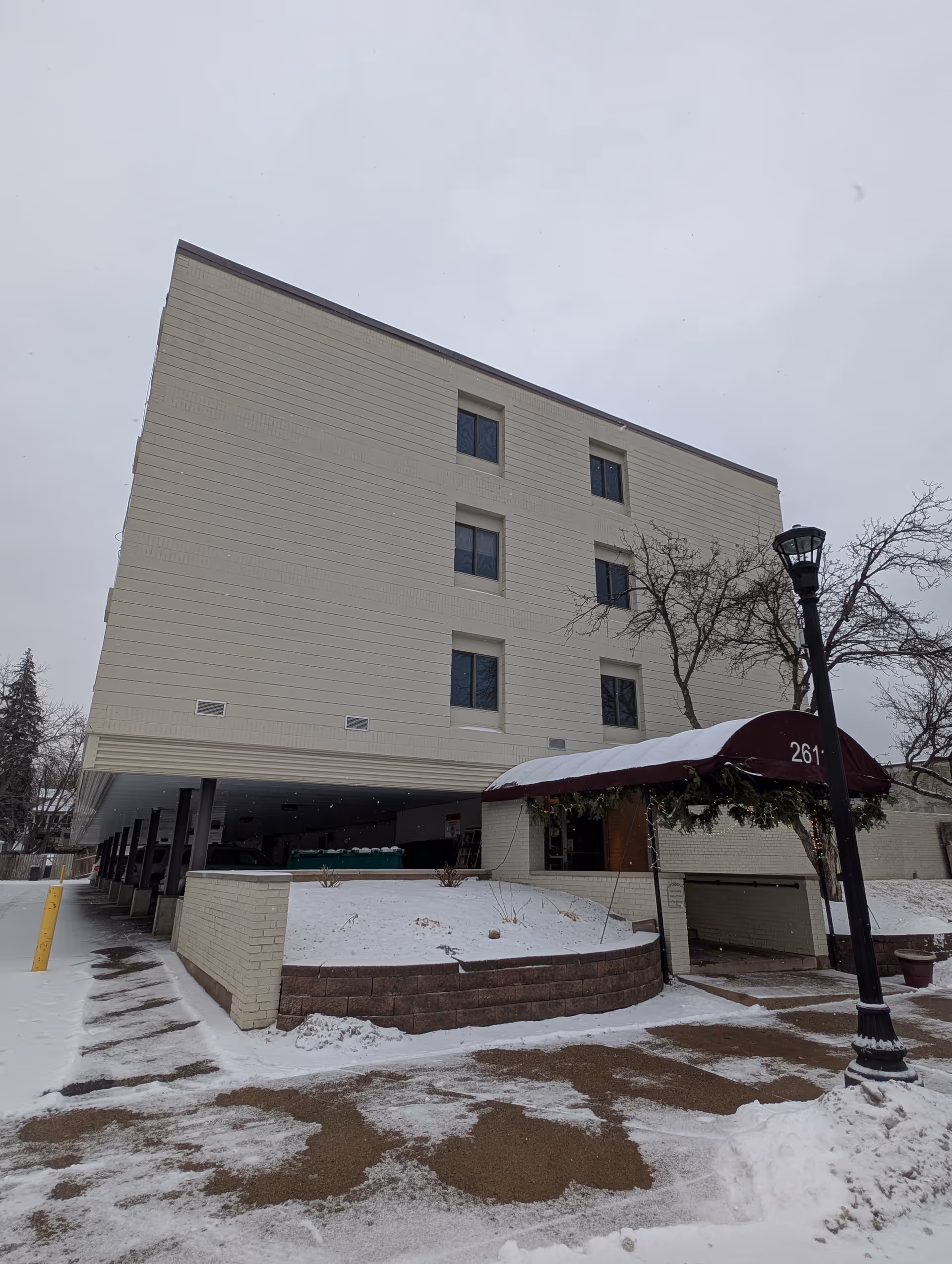 Snow-covered front exterior of a multi-story senior living building with a maroon awning over the entrance and a lamp post.