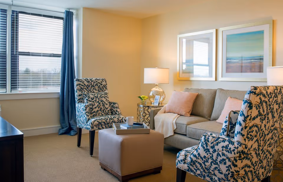 Sunlit living room with patterned armchairs, a beige sofa with pillows, an ottoman, side tables with lamps, and framed artwork.