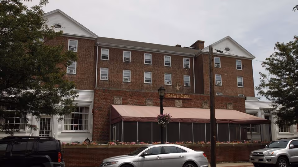 Exterior view of a multi-story brick building with white trim, identified as Madonna Manor Skilled Nursing & Rehabilitative Care. The building has multiple windows with air conditioning units, a covered patio area with a maroon awning, and several parked cars in front. Trees flank both sides of the building under an overcast sky.