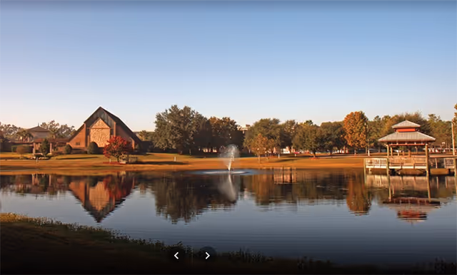 A calm lake with a central fountain reflecting trees, a gazebo on a dock, and buildings along the far shore under a clear sky.