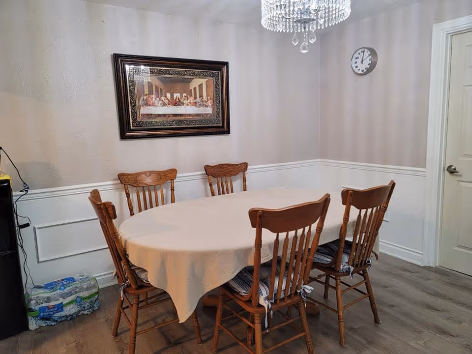 A dining room with a wooden oval table covered with a beige tablecloth and six wooden chairs with cushions. On the wall above the table is a framed picture of The Last Supper. A clock is mounted on the adjacent wall, and a chandelier hangs from the ceiling. There is a pack of bottled water on the floor near a black appliance.