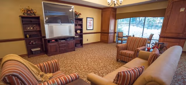A cozy living room area with patterned carpet, a large wooden entertainment center holding a television, two striped armchairs, a beige sofa with matching pillows, and a large window letting in natural light.
