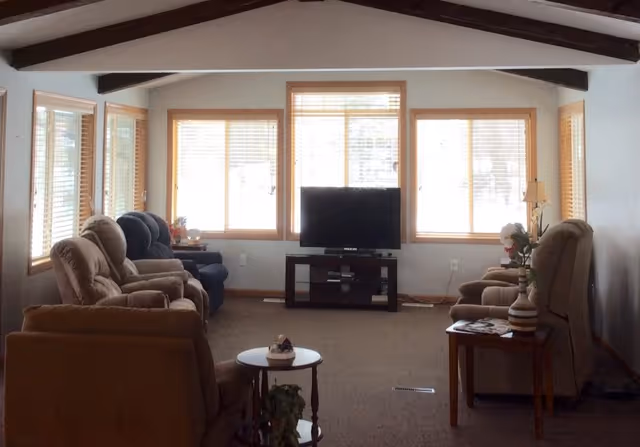 A cozy living room with multiple cushioned armchairs arranged facing a flat-screen TV on a black stand. The room has large windows with wooden frames and blinds, allowing natural light to brighten the space. There are small side tables with decorative items and a lamp, and exposed wooden beams on the ceiling.