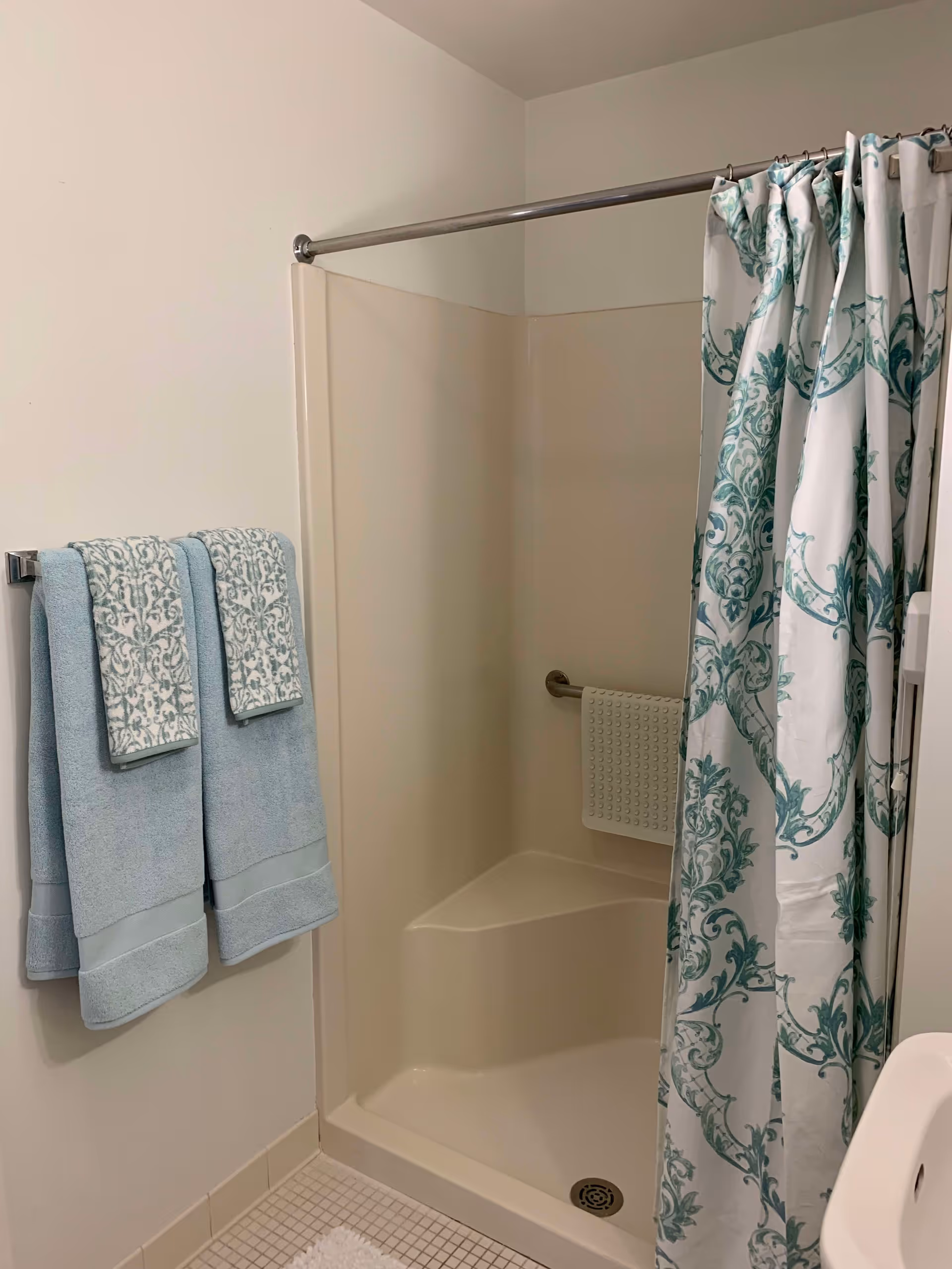 A bathroom shower area with a white and teal patterned shower curtain partially closed. There is a built-in corner seat inside the shower and a grab bar with a textured grip pad attached. Two light blue towels with patterned accents hang on a towel rack on the wall next to the shower. The floor has small square tiles and a white bath mat is partially visible.
