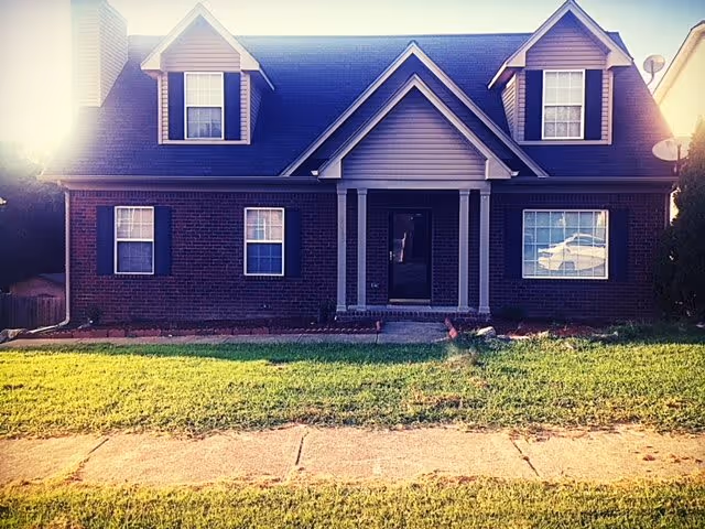 Front exterior view of a two-story brick house with a dark roof, two dormer windows, and a central entrance with a small porch. The house is surrounded by a green lawn and a sidewalk in front.