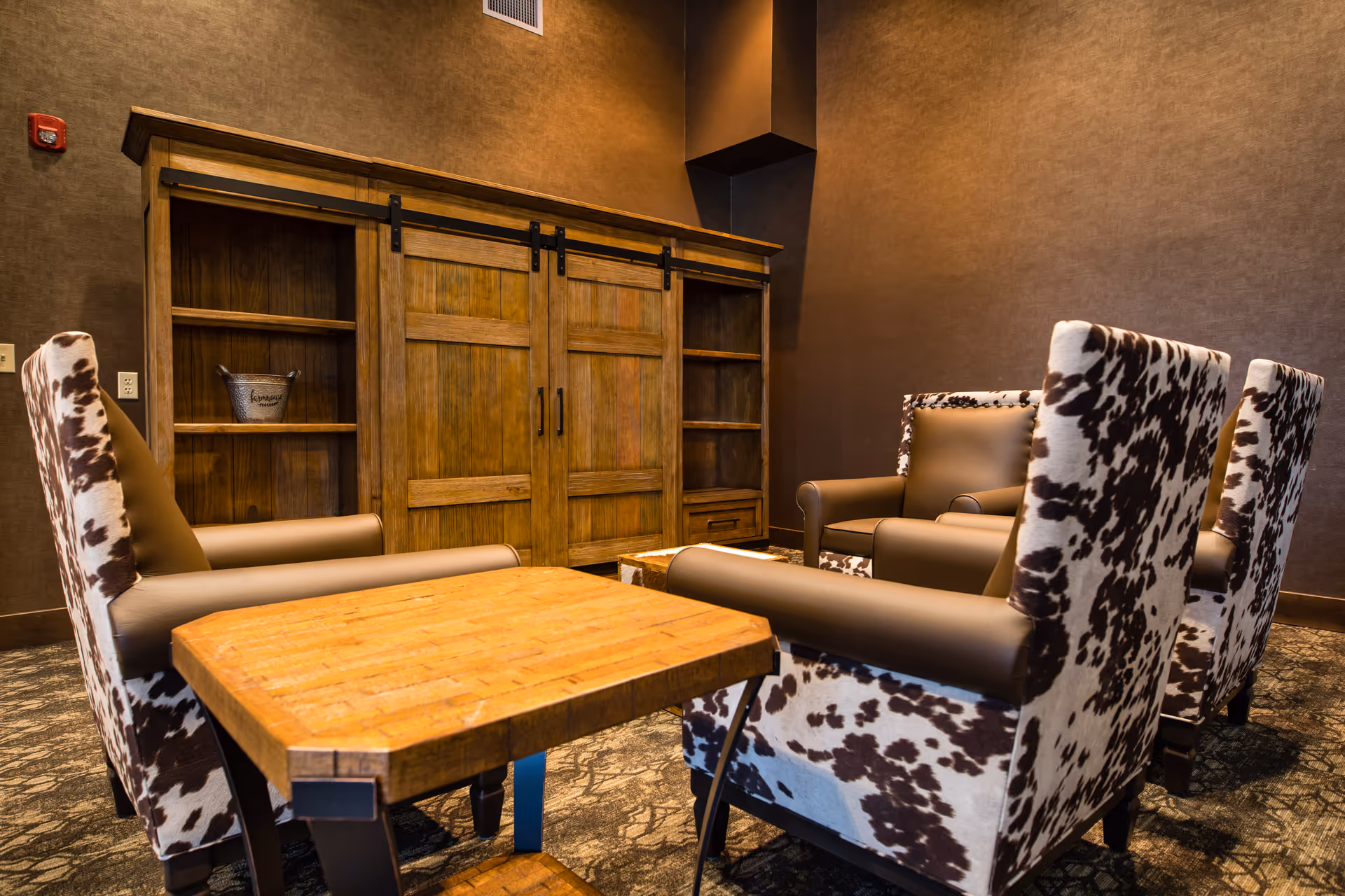 A cozy seating area with four armchairs upholstered in a brown and white cowhide pattern arranged around a wooden coffee table. Behind the chairs is a large wooden cabinet with sliding barn-style doors and open shelves on either side. The room has warm brown walls and carpeted flooring.