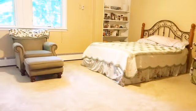 A cozy bedroom with a single bed featuring a decorative metal headboard and a floral bedspread. Next to the bed is a green armchair with an ottoman, positioned near a window with white trim. A built-in bookshelf filled with framed photos and decorative items is visible in the corner. The room has light-colored walls and carpeted flooring.