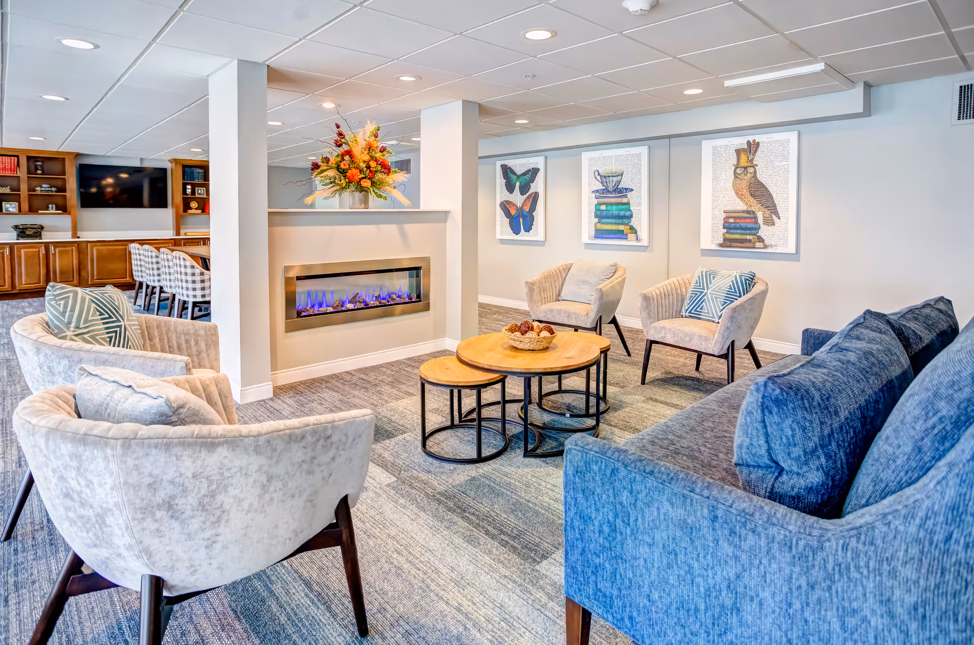 A cozy living room area in The Gardens Retirement Community featuring a modern electric fireplace with blue flames, surrounded by four beige armchairs and a blue sofa with patterned cushions. A set of three nested wooden coffee tables is in the center, with a decorative bowl on top. The walls are decorated with framed artwork of butterflies, a teacup on books, and an owl wearing a top hat and glasses. In the background, there is a wooden bookshelf with a TV and additional seating with plaid-upholstered chairs.
