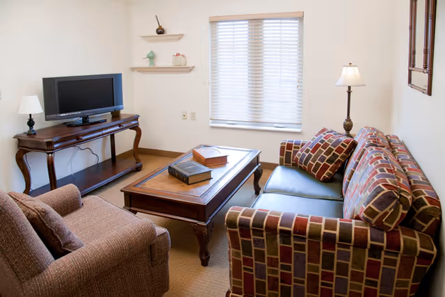 Cozy living room with a patterned sofa, armchair, wooden coffee table, TV on a console and a window with blinds.