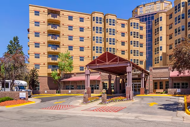 Exterior view of a multi-story senior living facility with a covered entrance driveway, landscaped areas with trees and flowers, and clear blue sky.