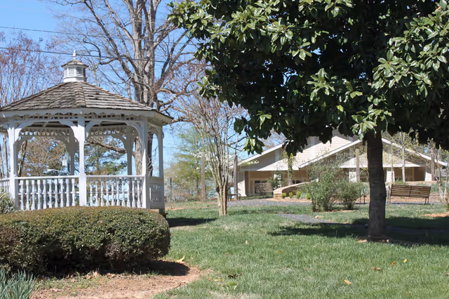 A white wooden gazebo with a shingled roof stands in a grassy area surrounded by bushes and trees. In the background, there is a beige building with benches outside, under a clear blue sky.
