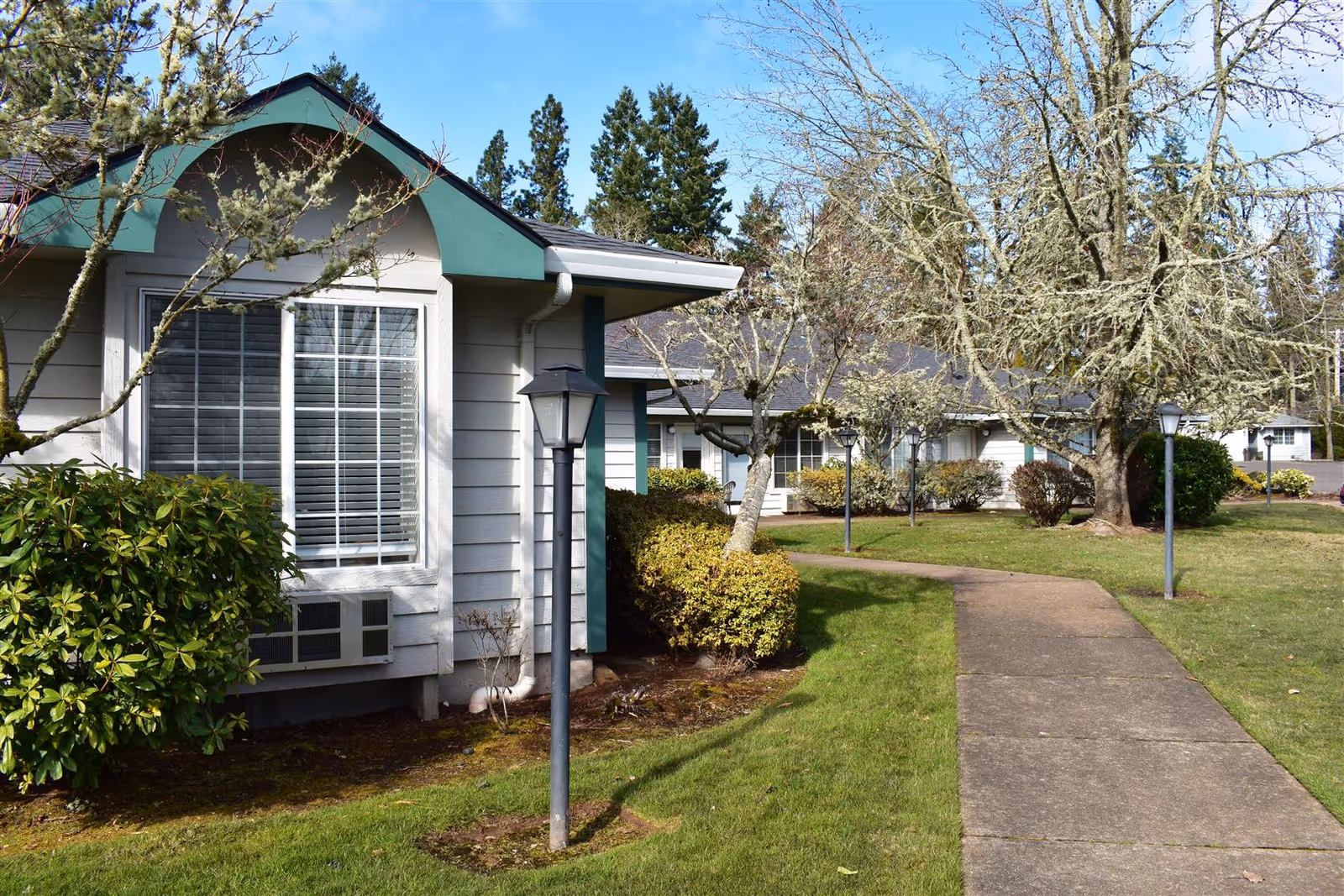 Exterior view of a senior living facility with a paved walkway, green grass, bushes, leafless trees, and single-story buildings with white siding and green trim under a clear blue sky.
