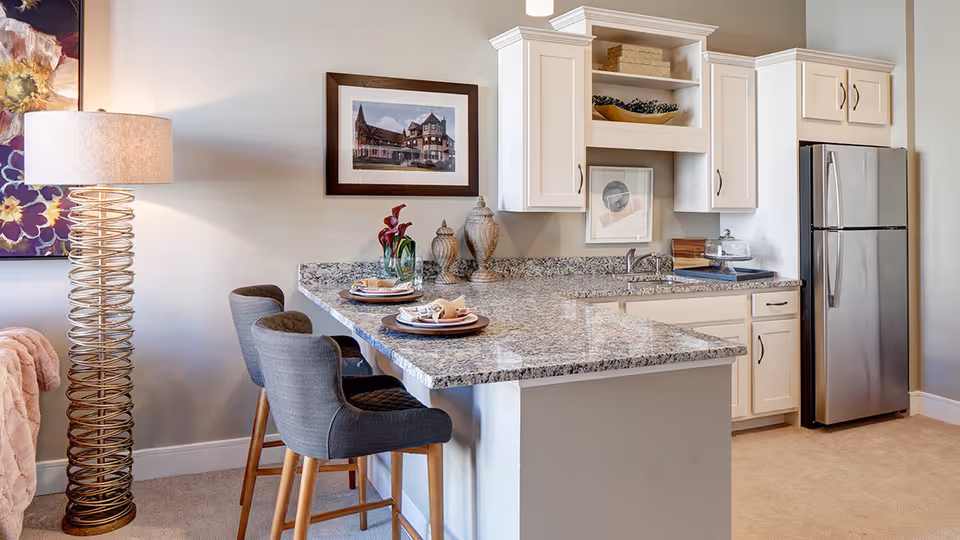 Open kitchen with a granite breakfast bar, two upholstered bar stools, white cabinets, and a stainless steel refrigerator.