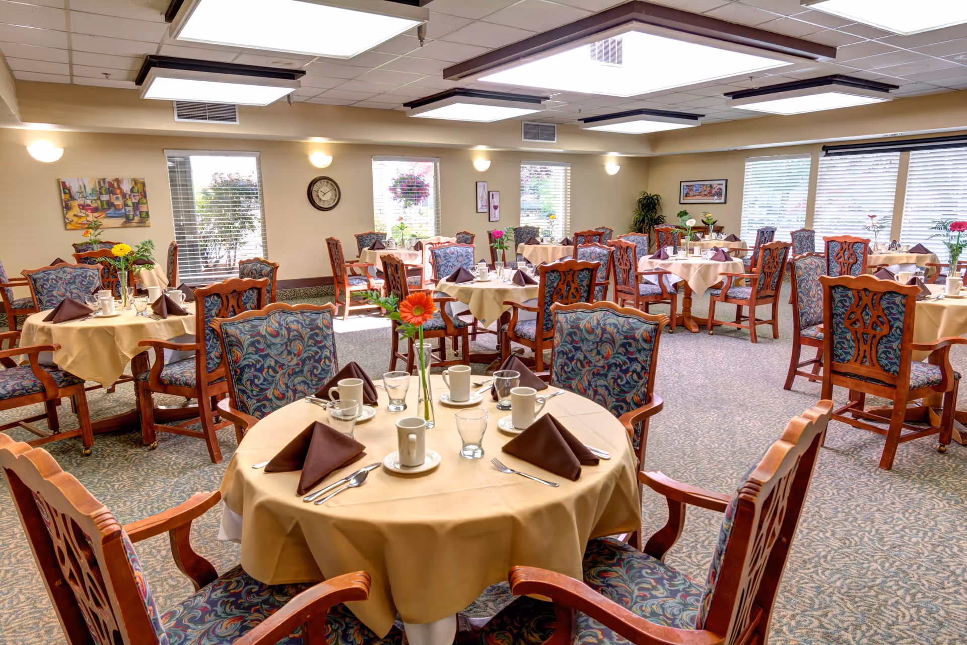 A bright dining room with multiple round tables covered with beige tablecloths, each set with brown folded napkins, cups, glasses, and silverware. The chairs have wooden frames with patterned upholstery. Large windows with blinds allow natural light to fill the room, and there are wall decorations including a clock and paintings.