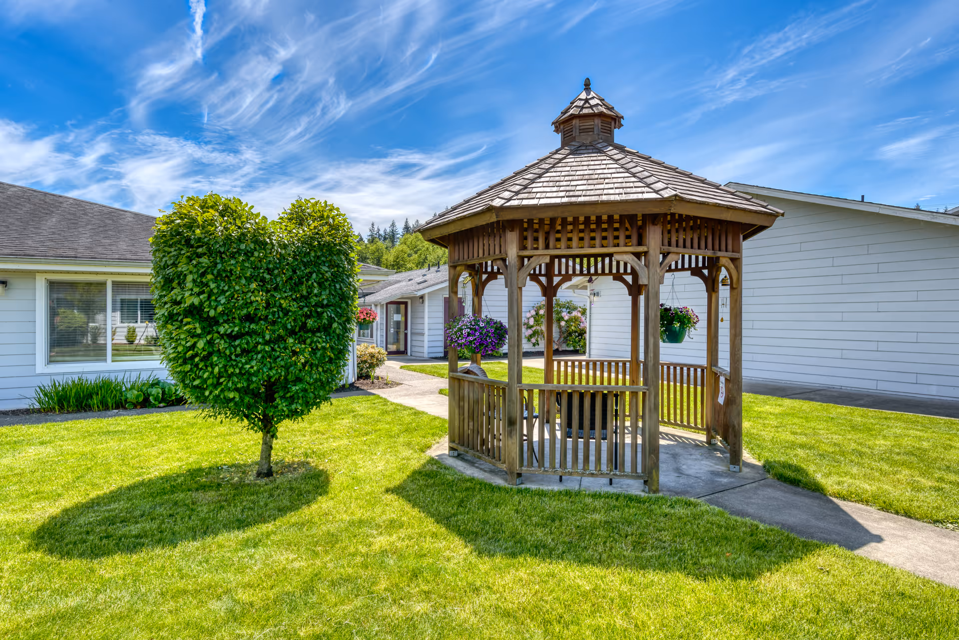 A well-maintained outdoor area at Beehive Retirement Community featuring a wooden gazebo with hanging flower baskets and a heart-shaped trimmed bush on a sunny day with a blue sky and wispy clouds.