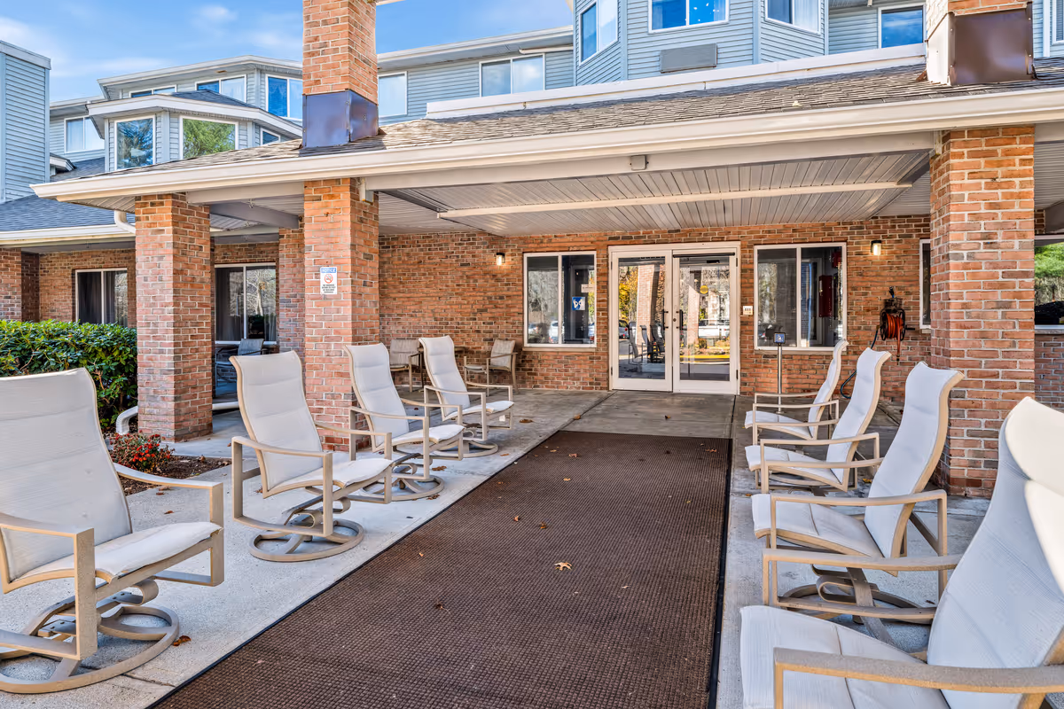 Covered outdoor seating area at the entrance of a senior living facility with cushioned swivel chairs arranged on both sides of a walkway leading to glass double doors. The building features brick pillars and siding with windows on either side of the entrance.