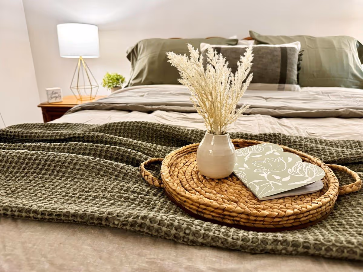 Close-up view of a neatly made bed with green and beige bedding, a woven tray holding a small white vase with dried flowers, and a floral-patterned notebook. A bedside table with a modern lamp and a small plant is visible in the background.