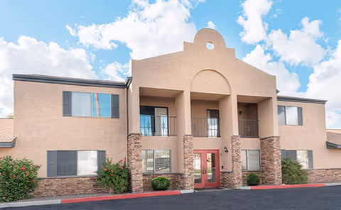 Front exterior of a two-story beige stucco building with a central arched entrance, balconies, and landscaping.