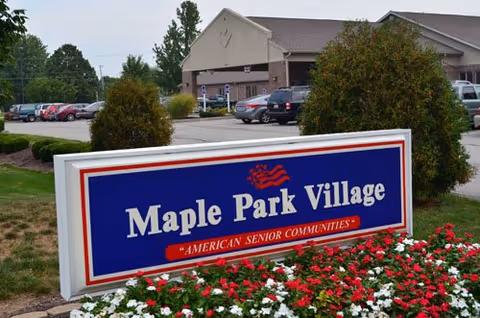 Outdoor sign reading "Maple Park Village" in front of flower beds with the facility building and parking lot in the background.