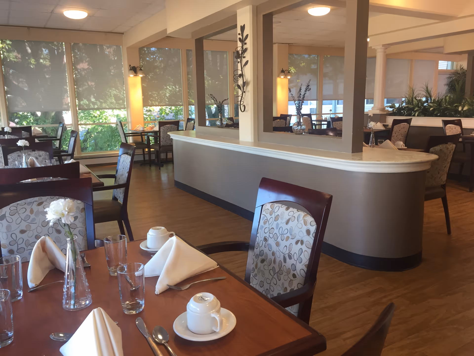 Well-lit dining room with set tables and chairs, folded napkins, glassware, and large windows overlooking greenery.