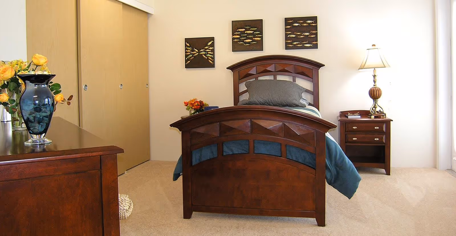 A bedroom with a wooden bed frame featuring a dark finish and geometric design. The bed is made with blue and gray bedding and a gray pillow. To the right of the bed is a wooden nightstand with a lamp and some small items. On the left side, there is a wooden dresser with a blue vase holding yellow flowers. The room has beige carpet and light-colored walls with three framed fish-themed artworks hanging above the bed. There are sliding closet doors in the background.