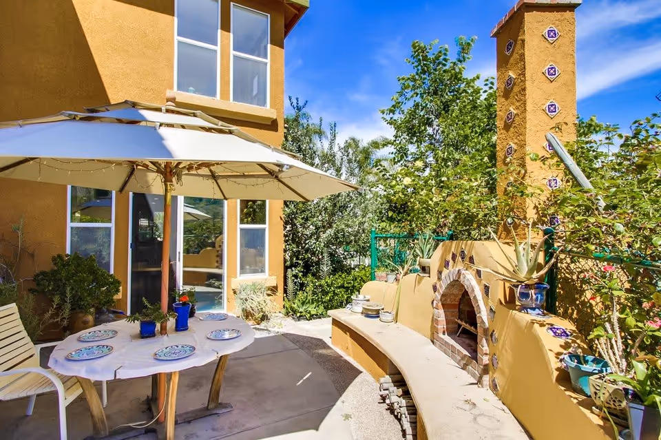 Outdoor patio area with a round table covered by a large umbrella, set with plates and cups. There is a built-in curved bench and a decorative outdoor fireplace with colorful tiles. The area is surrounded by greenery and plants, attached to a yellow stucco building under a clear blue sky.