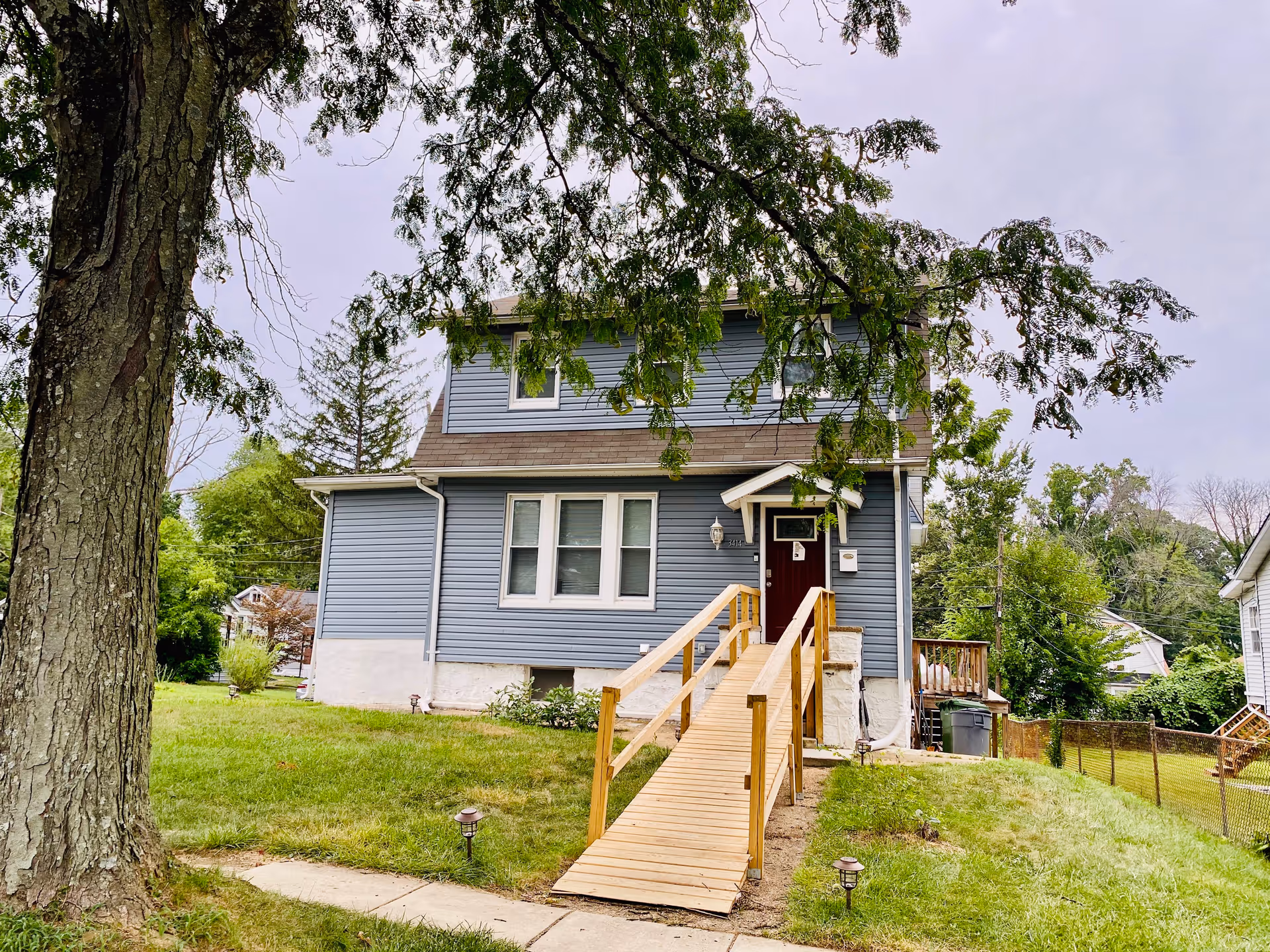 Exterior view of a two-story blue house with white trim and a brown roof. There is a wooden wheelchair ramp leading up to a dark red front door. The house is surrounded by green grass, trees, and neighboring houses in the background.