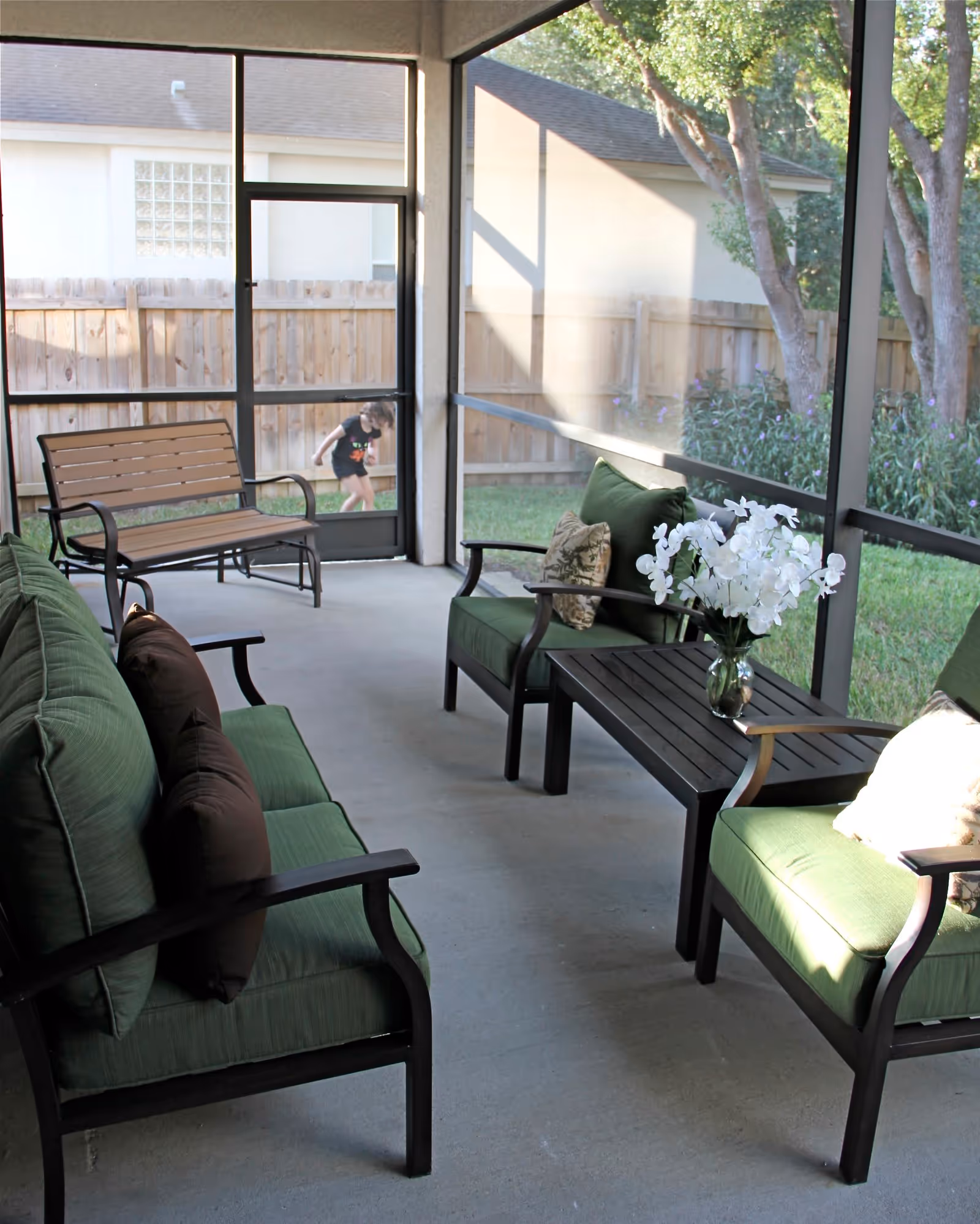 A screened-in porch with green cushioned chairs and a loveseat arranged around a black coffee table with a vase of white flowers. In the background, there is a wooden bench and a fenced yard with grass and trees.