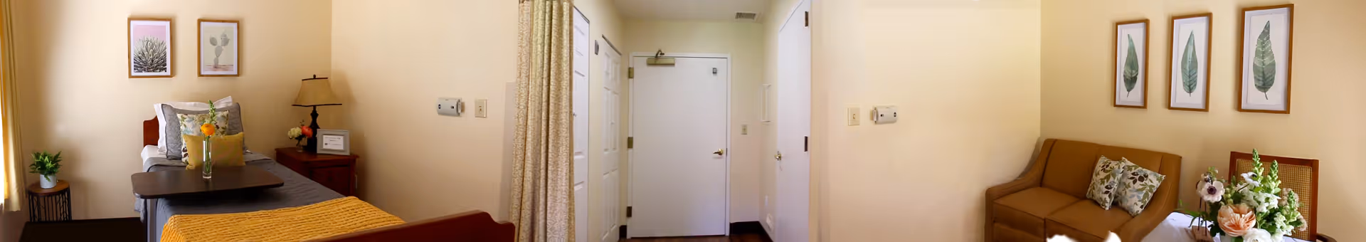 A cozy room in Villa Valencia Healthcare Center featuring a bed with a yellow blanket, a small side table with a lamp and framed pictures on the wall. There is a door in the center of the image, and on the right side, a brown couch with floral cushions and a small table with a flower arrangement.
